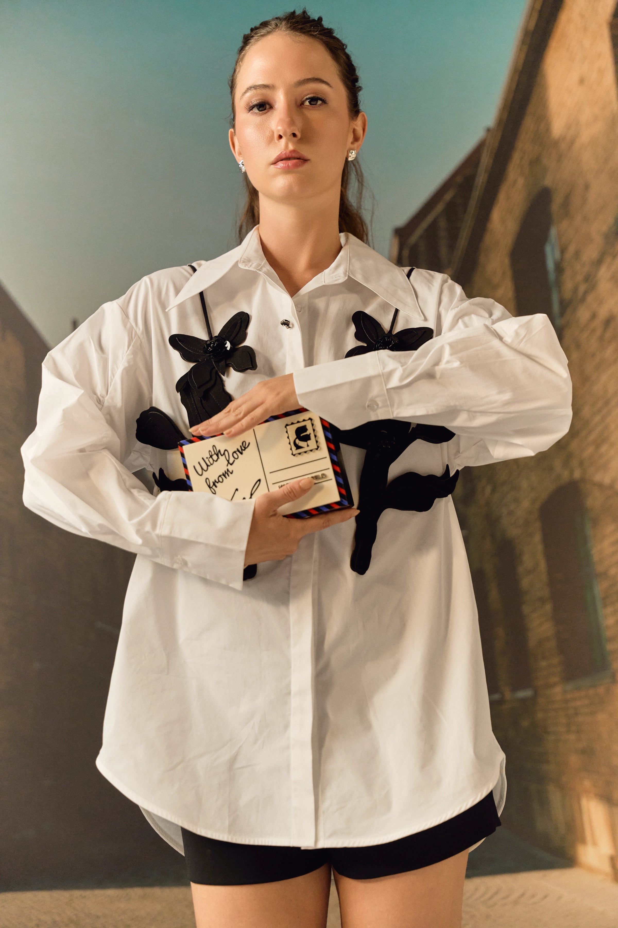 Person wearing a white shirt with black floral designs, holding a box, against an outdoor background.